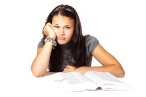 A teenager sitting with a book open in front of her, her head resting against one hand as she looks at the camera with a bored look on her face.