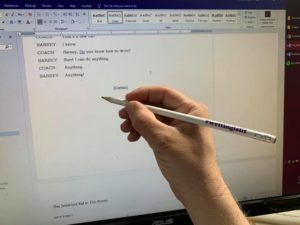 A hand holding a pencil that reads "Writing is Lit" up to a computer screen where a Word doc is showing a short play in progress.